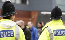 Two British policemen stand with backs to camera. Selectively focussed. A crowd of people appears in the background, out of focus. Policemen are wearing tall traditional British helmets and fluorescent Jackets.