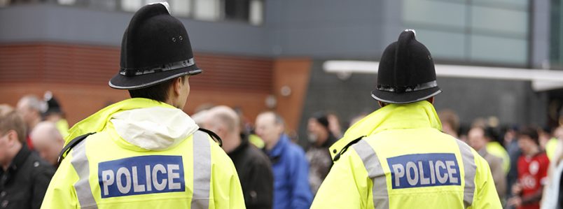 Two British policemen stand with backs to camera. Selectively focussed. A crowd of people appears in the background, out of focus. Policemen are wearing tall traditional British helmets and fluorescent Jackets.